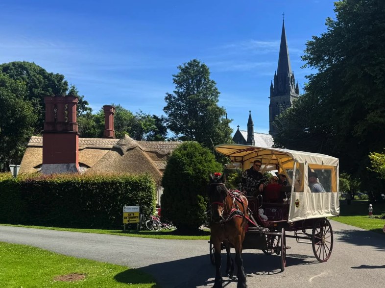 A covered jaunting car on the park drive past a thatched lodge, with a cathedral spire behind