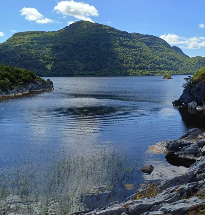 Still water and wooded shoreline on the Lakes of Killarney, below a hump of oak-covered mountain