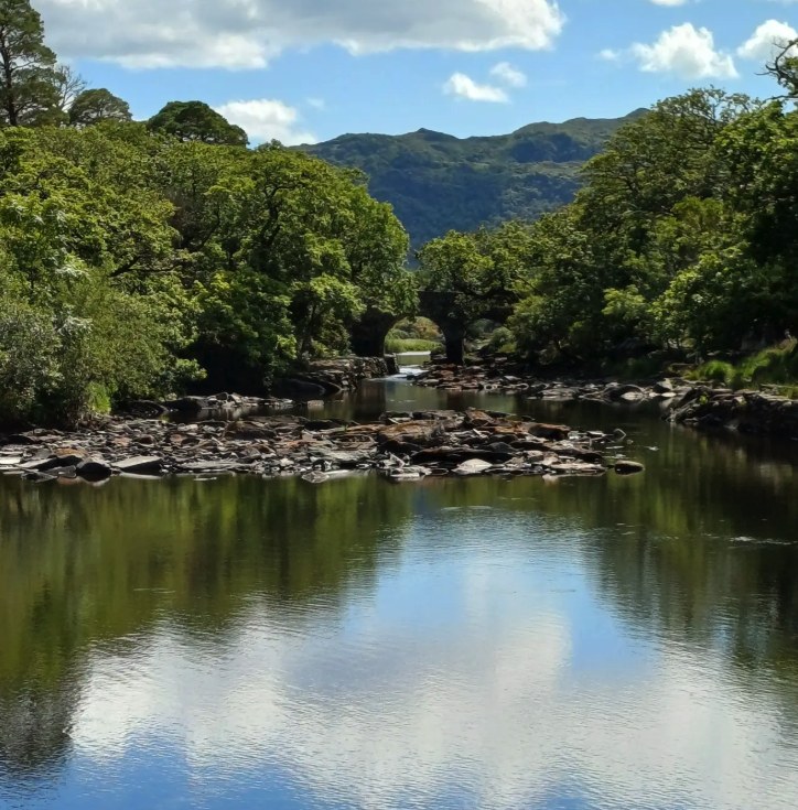 A stone arch bridge over the river between the Lakes of Killarney, with wooded hills behind